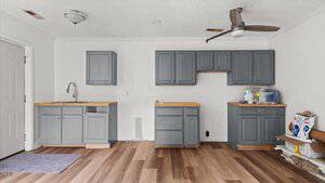 A small kitchen with gray cabinets, wood countertops, and no visible appliances. There is a ceiling fan, a sink on the left, and cleaning supplies on the right. The floor is wood, and the walls are white.