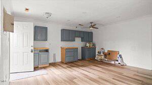 A partially renovated kitchen with unfinished gray cabinets, no appliances, exposed plywood, renovation supplies, and wood-look flooring. The walls are mostly white with visible patches, and the ceiling has a fan and light fixtures.