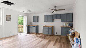 A mostly empty kitchen with gray cabinets, wooden countertops, a ceiling fan, a white door open to outside, and some cleaning supplies and materials on the counter and floor. No appliances are installed.
