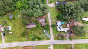 Aerial view of a rural property with several buildings, a driveway, trees, a round pool, parked vehicles, and power lines running parallel to a road at the bottom of the image.