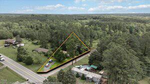 Aerial view of a triangular plot of land outlined in orange, bordered by a road, trees, and power lines, with a few buildings and vehicles nearby and woods in the background.