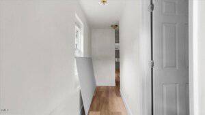 A narrow hallway with white walls and light wood flooring, a window on the left, an interior door on the right, and a large rectangular board leaning against the wall. The hallway leads to a kitchen area.