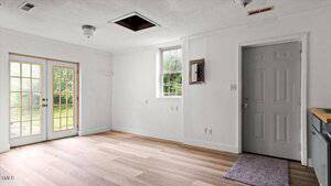 An empty, unfurnished room with light wood floors, white walls, a closed gray door, a window, French doors leading outside, a small area rug, and an open ceiling hatch.