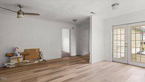 A mostly empty room with light wood flooring, white walls, a ceiling fan, a pile of construction materials on the floor, and double glass doors leading outside. A hallway and another room are visible in the background.
