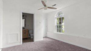 A small, unfurnished room with light carpeting, white walls, a ceiling fan, an arched window, and an open doorway leading to a bathroom with a toilet and vanity.