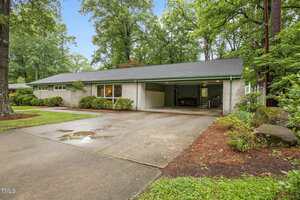 A single-story house with light brick exterior, gray roof, two-car carport, and neatly trimmed bushes, surrounded by tall trees and a wet concrete driveway on a cloudy day.