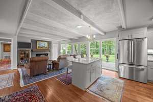 Open-concept living area with a kitchen island in the foreground, adjacent to a seating area with armchairs, a fireplace, bookshelves, and large windows overlooking a patio and green yard.