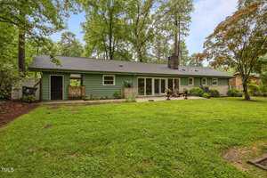 Single-story house with green siding, multiple windows and glass doors along the back, a small patio seating area, and a large grassy backyard bordered by trees and shrubs.