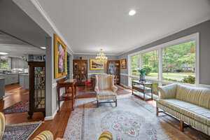A living room with large windows, patterned armchairs, a cream sofa, wood furniture, artwork on the walls, a chandelier, and an ornate rug, with a view into a kitchen and dining area.