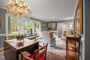 A living room with large windows, gray walls, a chandelier, armchairs, framed paintings, an area rug, a desk with chairs, and a side table with a lamp. Trees and houses are visible outside.