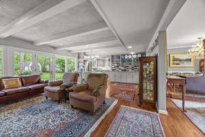 A spacious living area with large windows, brown leather sofa, two striped armchairs, multiple rugs, hardwood floors, open kitchen with white cabinets, and a dining area with a chandelier and piano.