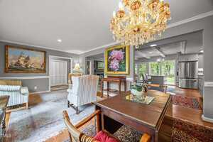 A well-lit living and dining area features a crystal chandelier, traditional furniture, framed paintings, hardwood floors with rugs, and visible kitchen appliances in the background. Large windows offer a view of greenery outside.