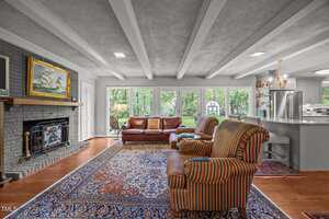 A living room with a red leather sofa, two striped armchairs, a patterned rug, a fireplace with a painting above it, and large windows overlooking a backyard; an open kitchen is visible to the right.