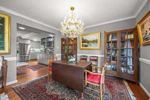 A dining room with a dark wood table, four chairs with red cushions, glass-front cabinets, a crystal chandelier, and framed paintings. The room connects to a kitchen with stainless steel appliances and patterned rugs on hardwood floors.