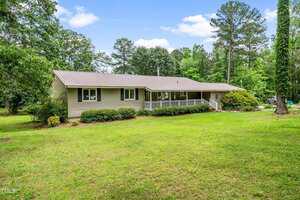 Charming single-story beige house at 608 20th Street, Butner, with a metal roof and front porch, nestled among green grass, shrubs, and tall trees under a partly cloudy sky.