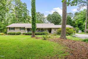 A single-story house with beige siding and a metal roof sits behind a green lawn with scattered trees and shrubs. A driveway is visible on the right, and the area is surrounded by tall, leafy trees.
