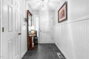 A small entryway with white walls and wainscoting, black tile flooring, a white front door, a side table with a lamp and decor, a framed photo on the wall, and a ceiling light fixture.