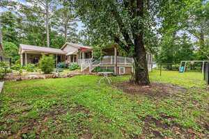 A single-story house with a metal roof and covered porch, surrounded by trees and grass. There is a white metal table and chairs on the lawn and a black chain-link fence in the backyard.