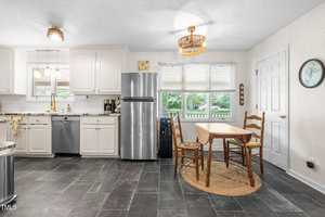 A kitchen with white cabinets, stainless steel appliances, and black tile flooring. There is a wooden dining table with two chairs on a circular rug near a window, and a wall clock is mounted on the right wall.