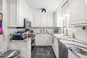 A modern kitchen with white cabinets, stainless steel appliances, a marble-patterned countertop, a double sink, and black tiled floors. Pendant lights hang over the sink, and small appliances are on the counters.