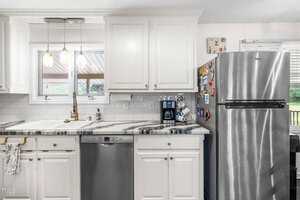 A kitchen with white cabinets, a marble countertop, stainless steel dishwasher, and refrigerator. A sink is under a window, and various magnets and items are on the fridge. Pendant lights hang above the sink.