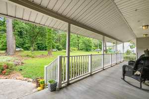 A covered front porch with white railing, gray flooring, and black wicker chairs overlooks a green lawn with trees and bushes. Potted plants and yellow rain boots are placed near the porch steps.
