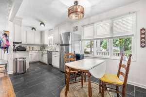 A bright kitchen with white cabinets, stainless steel appliances, a small dining table with two wooden chairs, black tile floor, and large window letting in natural light.