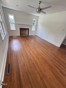 A bright, empty room with hardwood floors, white walls, a ceiling fan, several windows, and a white fireplace with a red-brick interior. The room is well-lit with natural light.