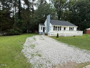 Single-story white house with a dark roof, a front porch with railing, surrounded by trees, and a gravel driveway leading from the street to the house on a grassy lawn.