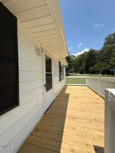 A wooden front porch with white railing attached to a white house. The door has a window, and there is a light fixture beside it. Trees and a lawn are visible in the background on a sunny day.