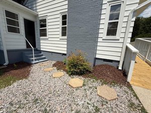 A small shrub is planted along a gray gravel path with hexagonal stepping stones beside a white house. The house has gray brick accents, a set of stairs, and a wooden ramp leading to the entrance.