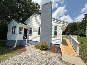 Single-story white house with a gray brick chimney, small front porch with steps, a wooden ramp for accessibility, gravel walkway, and grassy yard with trees in the background under a partly cloudy sky.