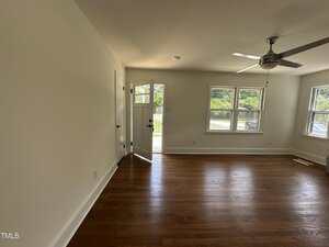 A room with hardwood floors, white walls, a ceiling fan, three large windows, and a door that is slightly open to the outside. Sunlight streams in, illuminating the space.