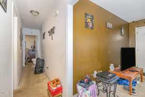 A hallway with beige carpet leads to a room with a brown accent wall. Shoes and personal items are on the floor, and a TV sits on a stand. Framed wall art decorates the walls. The space appears cluttered.