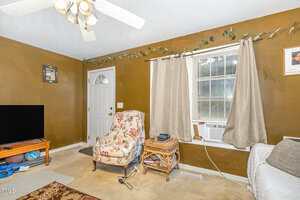 A living room with brown walls, a ceiling fan, a window with beige curtains and an air conditioner, a floral armchair, a wicker side table, a TV on a stand, and a closed white door.