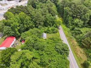 Aerial view of a rural area with a paved road running through dense green trees, a white building with a red roof on the left, and smaller structures partially hidden by foliage. Industrial buildings are visible in the top left corner.