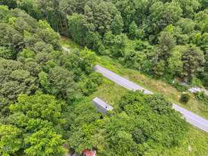 A paved road runs through a densely wooded, green area. A small building with a gray roof is partially visible near the road, surrounded by trees and vegetation.