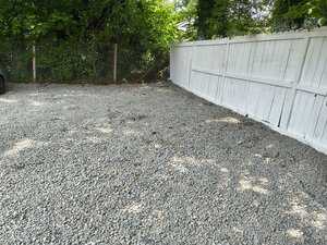 A gravel parking area bordered by a white wooden fence on the right and a chain-link fence with green foliage on the left and background. A portion of a parked vehicle is partially visible on the left edge.