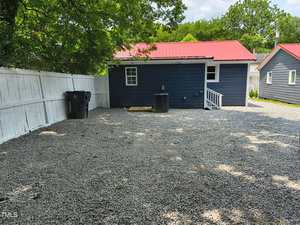 A small dark blue house with a red roof sits behind a gravel yard, bordered by a white wooden fence. There are two trash bins, an air conditioning unit, and a few trees providing shade.