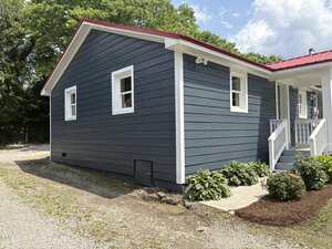 A small, single-story house with dark blue siding, white trim, and a red metal roof. There are plants and mulch along the foundation, a concrete walkway, and a gravel driveway beside the house.