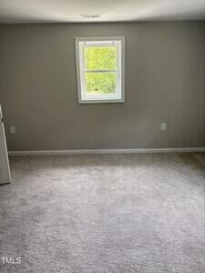A small, empty room with light gray walls, a single white-framed window, beige carpet, and two electrical outlets visible on the walls. Sunlight is coming through the window.
