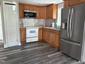 A kitchen with wooden cabinets, white stove and microwave, stainless steel refrigerator, gray countertops, tile backsplash, sink with dish soap, and gray wood-look flooring.