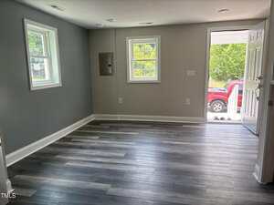 An empty room with gray walls, two windows, wood-look flooring, and an open white door leading outside. A red vehicle is partially visible through the door. An electrical panel is on one wall.