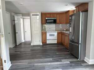 A kitchen with wood cabinets, gray tile backsplash, white appliances, stainless steel refrigerator, and gray wood-look flooring. Two white doors are visible on the left side of the image.