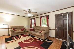 A living room with two beige sofas, a patterned area rug, a floor lamp, two windows with red curtains, and a dark wooden front door. The floor is hardwood and a ceiling fan with a light is overhead.