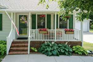 A small house with green shutters and a white porch, featuring red flowers in boxes and hanging baskets, an American flag, brick steps, and leafy green plants along the front walkway.