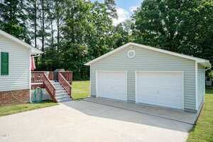 A two-car detached garage with white doors and light gray siding sits next to a house with green shutters and a red staircase, surrounded by trees and a concrete driveway.