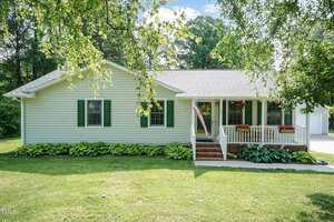 Single-story house with light green siding, green shutters, and a covered front porch. An American flag hangs by the steps, with flower boxes on the porch railing and a neatly maintained lawn in front.