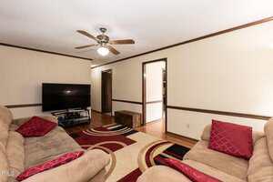 A living room with beige walls and wood trim, two tan sofas with red pillows, a patterned area rug, a flat-screen TV on a stand, a ceiling fan with lights, and hardwood flooring.