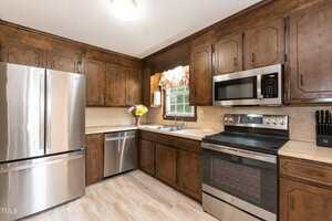 A kitchen with dark wooden cabinets, stainless steel appliances including a refrigerator, dishwasher, oven, and microwave, a sink under a window with a floral curtain, and a knife block on the counter.